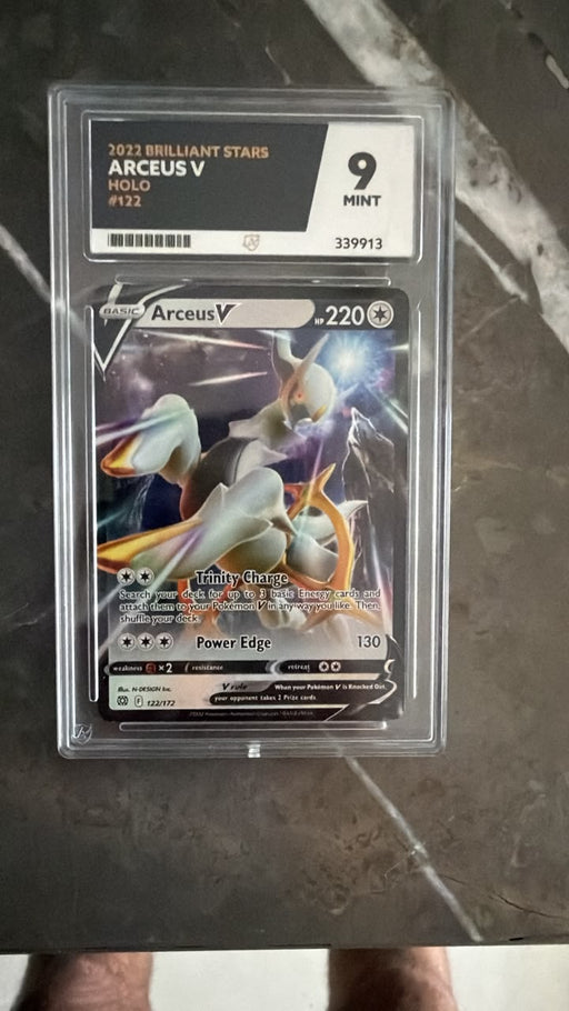 A graded Pokémon TCG card, Arceus V Holo #132 from the 2022 Brilliant Stars set, sealed in a clear PSA-style holder and photographed on a dark marble-like surface.
